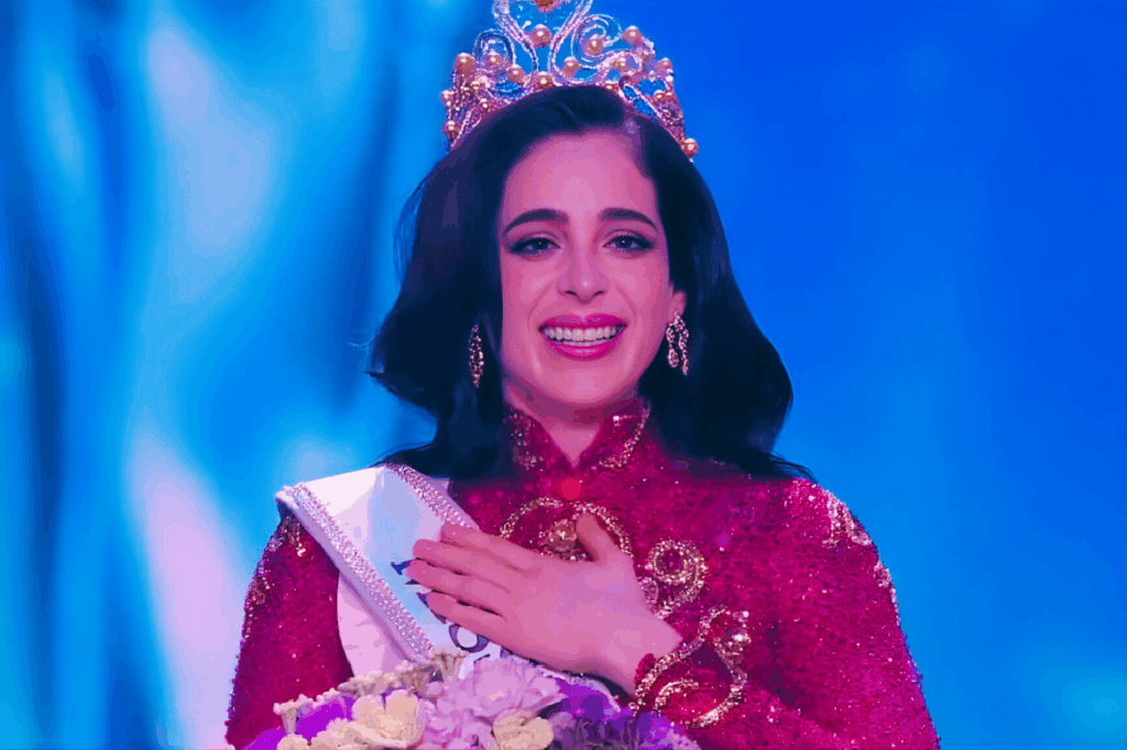 Mujer coronada con traje rojo y corona dorada, sonriendo emocionada en el escenario de Miss Universe 2025.