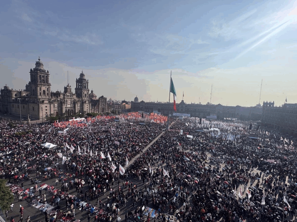 Vista aérea del Zócalo capitalino completamente lleno de contingentes con banderas y colores de Morena durante el mitin por los siete años del triunfo de la Cuarta Transformación, con la Catedral Metropolitana y la bandera monumental al fondo.