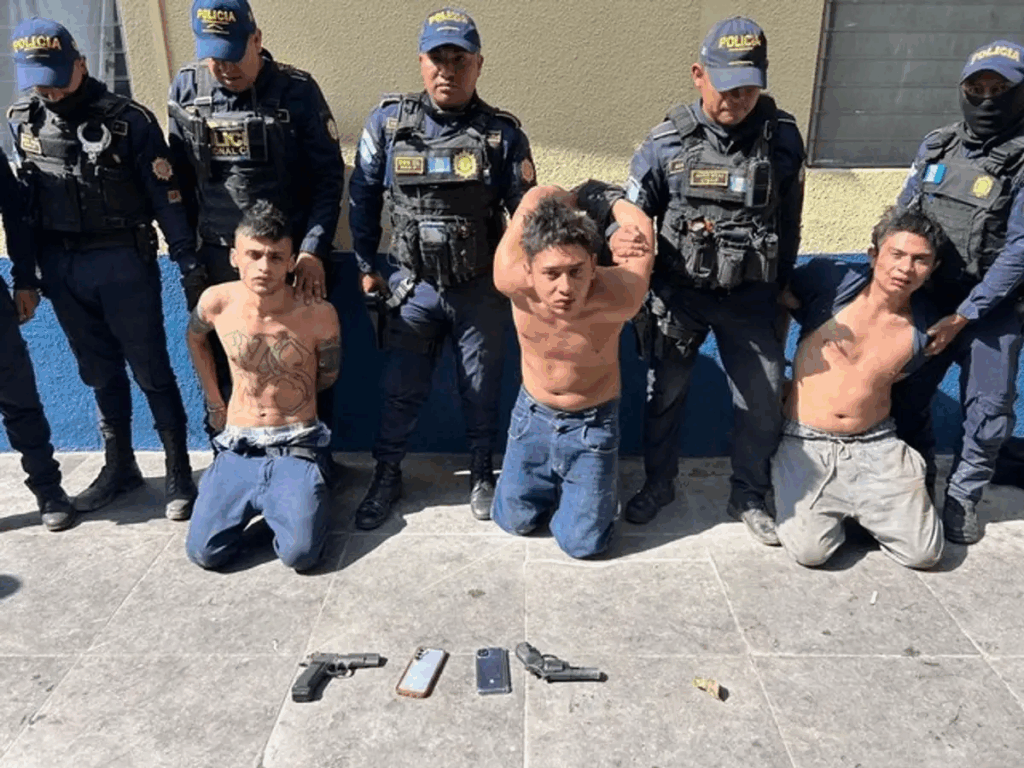 Agentes de la Policía Nacional Civil custodian a tres presuntos integrantes de pandillas detenidos durante operativos de seguridad en Guatemala; al frente se observan armas cortas y teléfonos asegurados como evidencia.