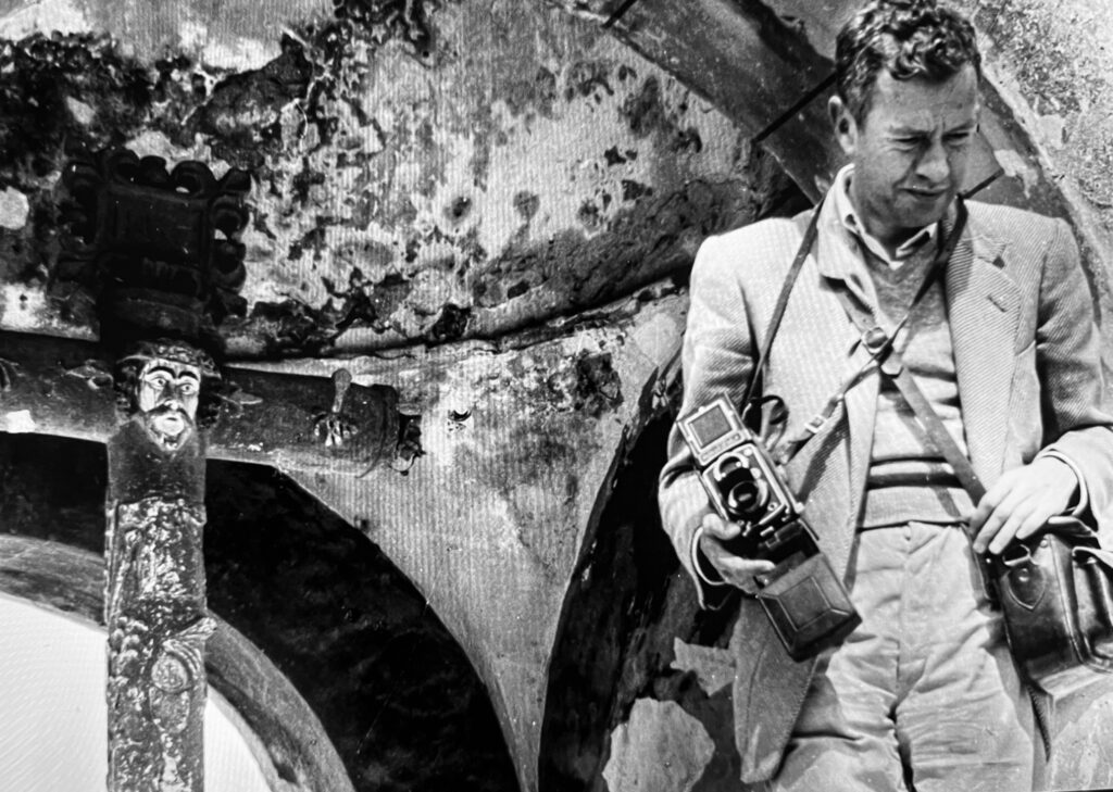 Escritor mexicano del siglo XX sostiene una cámara fotográfica frente a una construcción antigua.