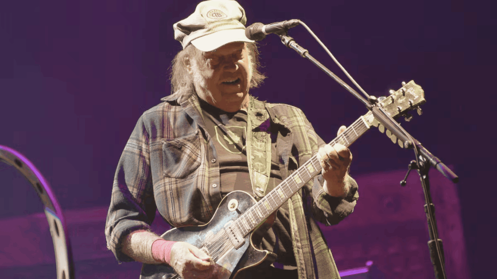 Neil Young tocando guitarra eléctrica en vivo durante un concierto, bajo luces moradas de escenario.