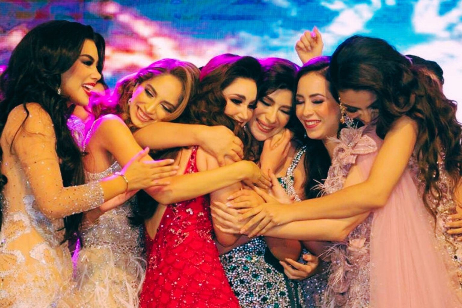 Fotografía moderna y vibrante de un abrazo grupal en el escenario de la Feria Tabasco. Varias jóvenes candidatas del certamen Flor de Oro, vestidas con trajes de noche de gala brillantes y bordados, se abrazan eufóricas. Se aprecian los detalles del confeti y la audiencia tomando fotos. Estilo moderno con marco redondeado.